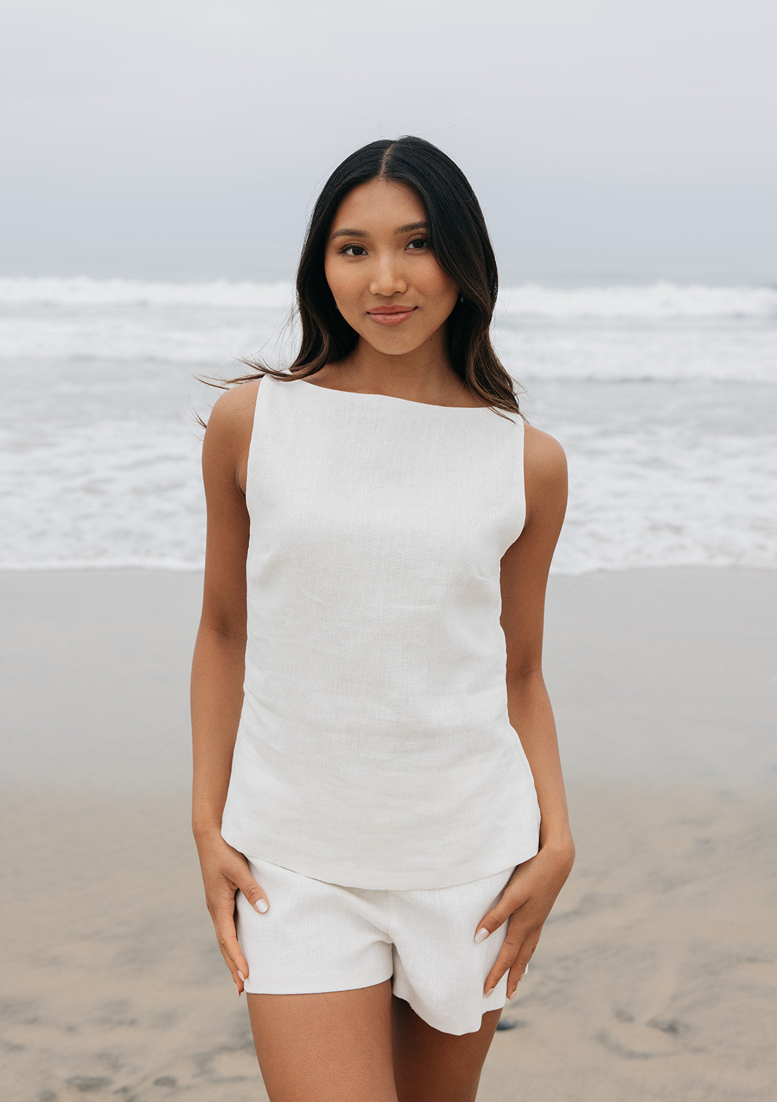 Woman in a white sleeveless top and shorts standing on a beach.