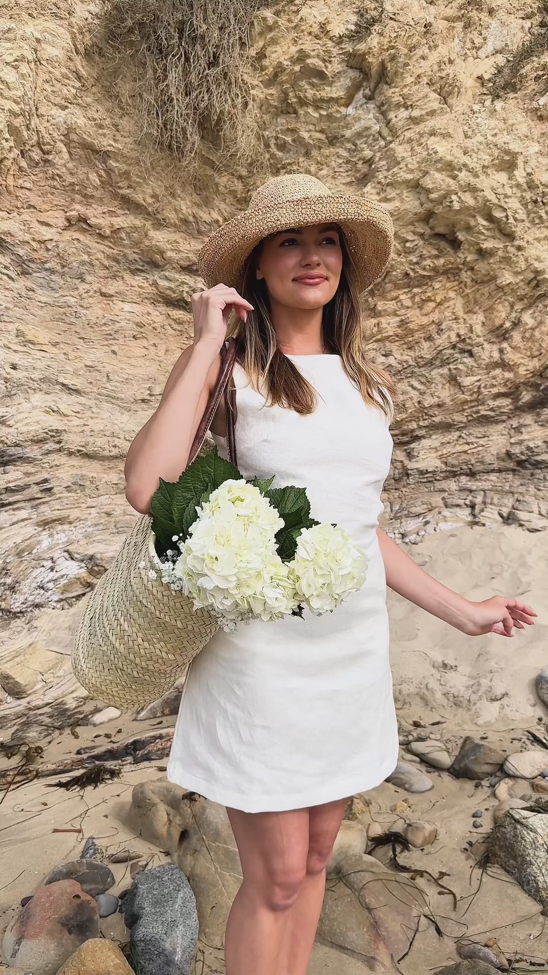 Model wearing Bardot Shift Dress in white | linen | styled with hat, basket bag and gold jewelry | at the ocean