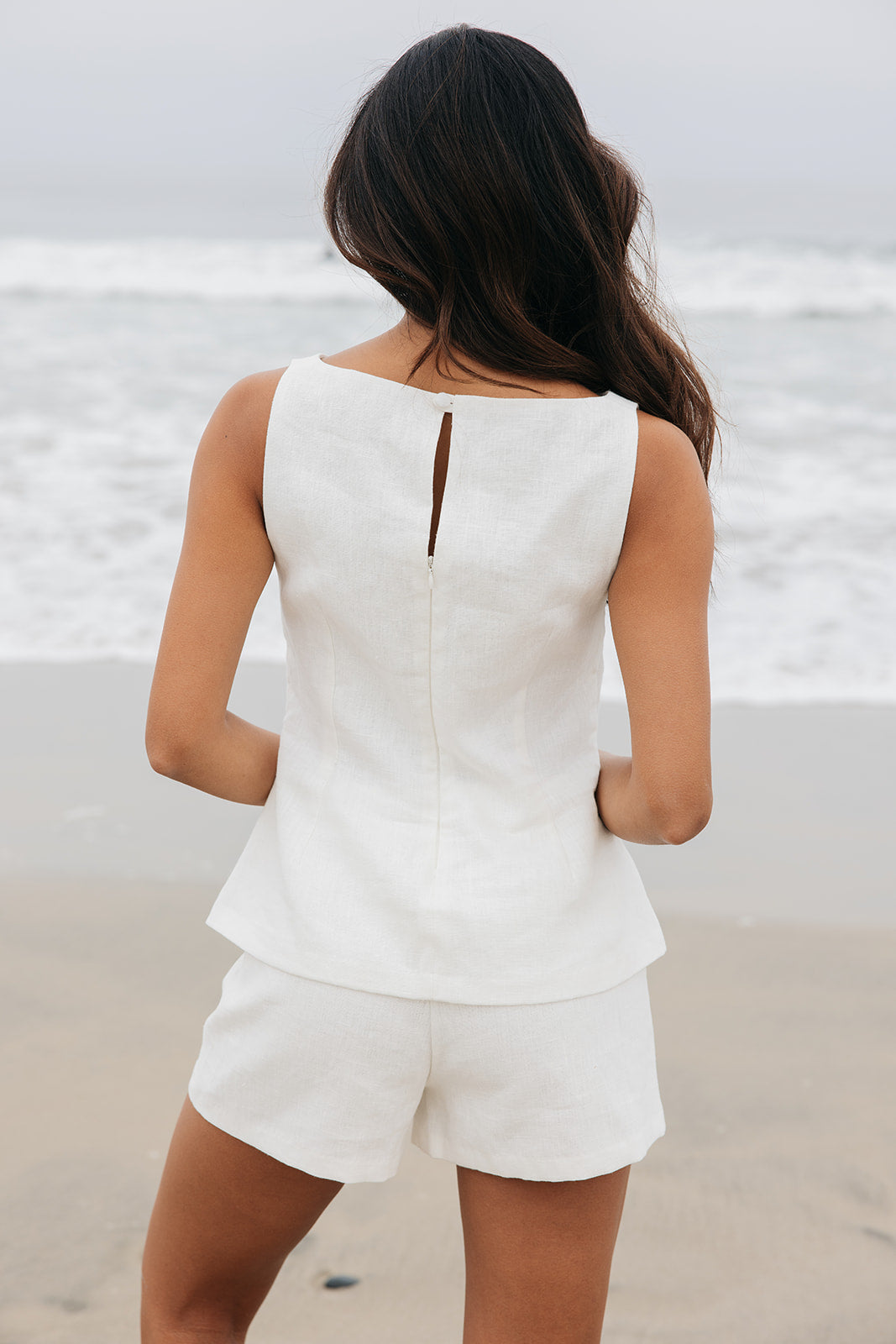 Woman wearing a white sleeveless top and shorts on a beach.